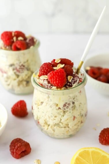 A jar of raspberry lemon overnight oats topped with extra raspberries and lemon zest. A spoon rests in the jar. A second jar is in the background.