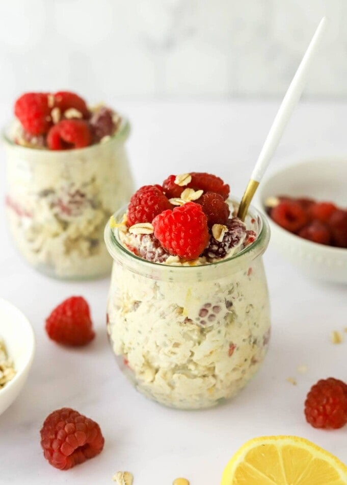 A jar of raspberry lemon overnight oats topped with extra raspberries and lemon zest. A spoon rests in the jar. A second jar is in the background.