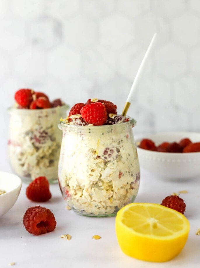 A jar of raspberry lemon overnight oats topped with extra raspberries and lemon zest. A spoon rests in the jar and a second jar is in the background out of focus.
