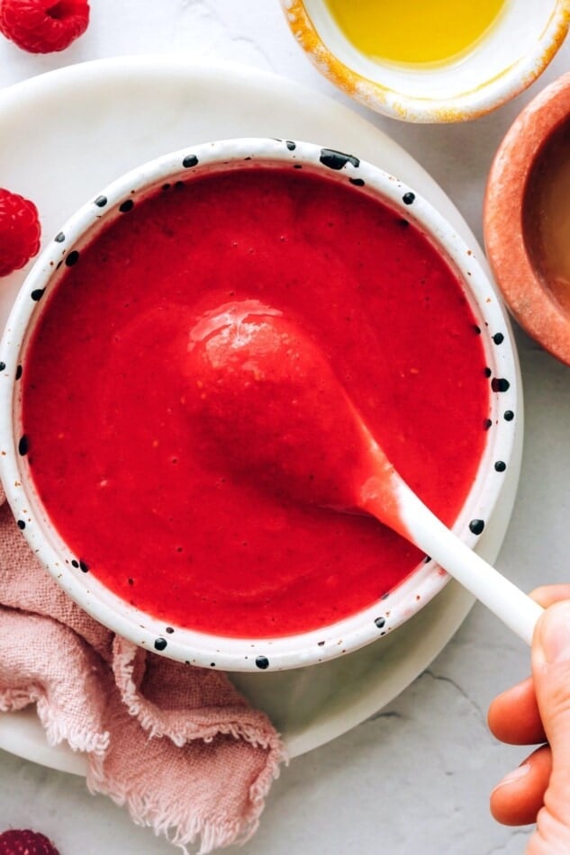 A small dish holding raspberry vinaigrette. A small spoon is lifting some dressing out of the bowl.