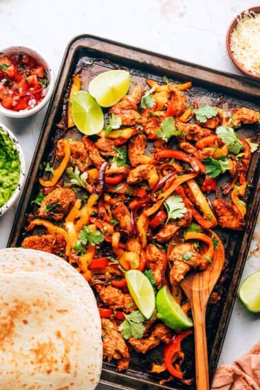 Chicken fajita filling on a sheet pan with a wooden spoon. Tortillas are resting on the bottom of the sheet pan.