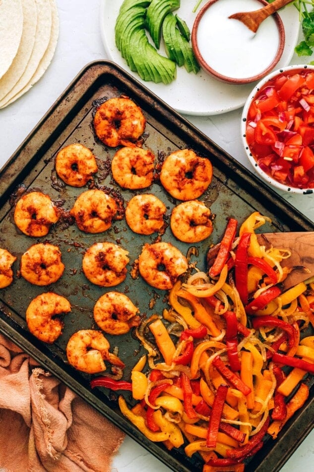Cooked veggies and shrimp on a sheet pan. Fajitas accoutrements surround the sheet pan.