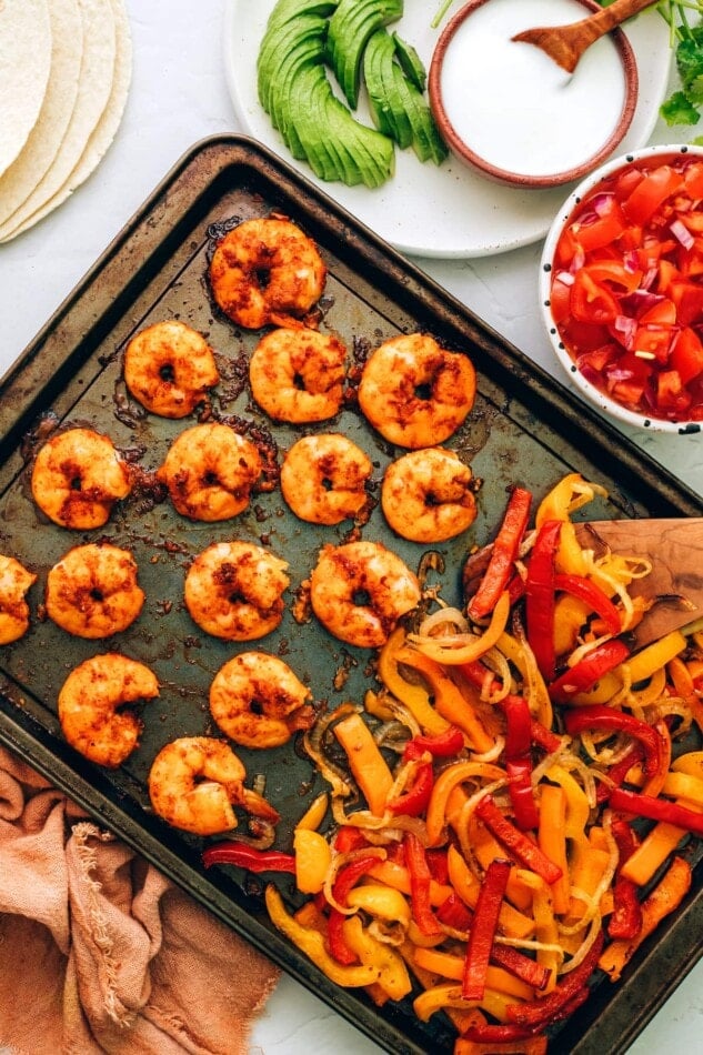 Cooked veggies and shrimp on a sheet pan. Fajitas accoutrements surround the sheet pan.