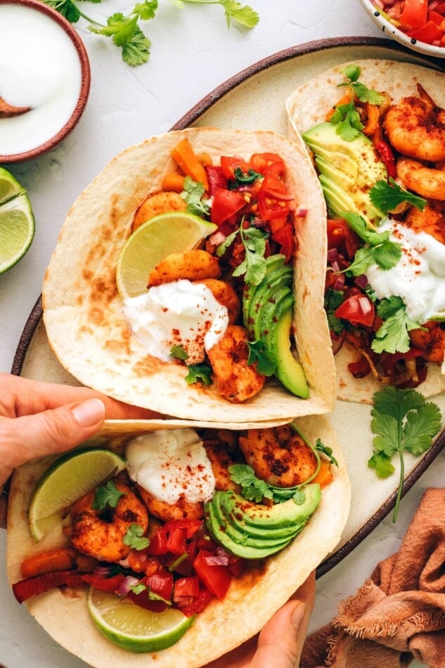 Three fajitas on a serving plate. Hands are removing a fajita.