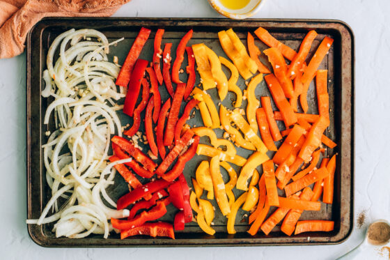 Sliced onion, red pepper, yellow pepper and orange pepper on a sheet pan.