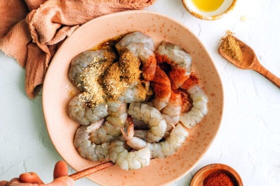 Raw shrimp being tossed with olive oil, chili powder, cumin, onion powder and salt.