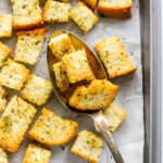 Sourdough croutons on a baking sheet lined with parchment paper. A spoon is lifting a few croutons off the baking sheet.