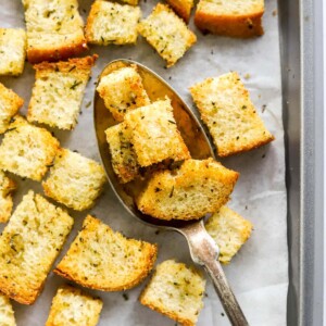 Sourdough croutons on a baking sheet lined with parchment paper. A spoon is lifting a few croutons off the baking sheet.