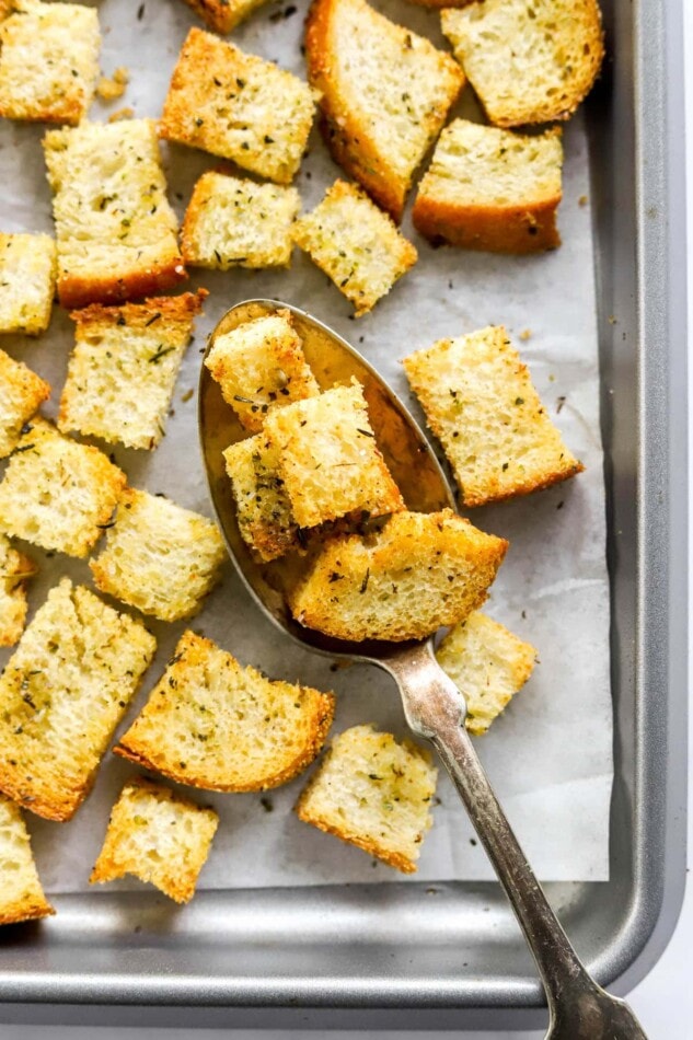Sourdough croutons on a baking sheet lined with parchment paper. A spoon is lifting a few croutons off the baking sheet.