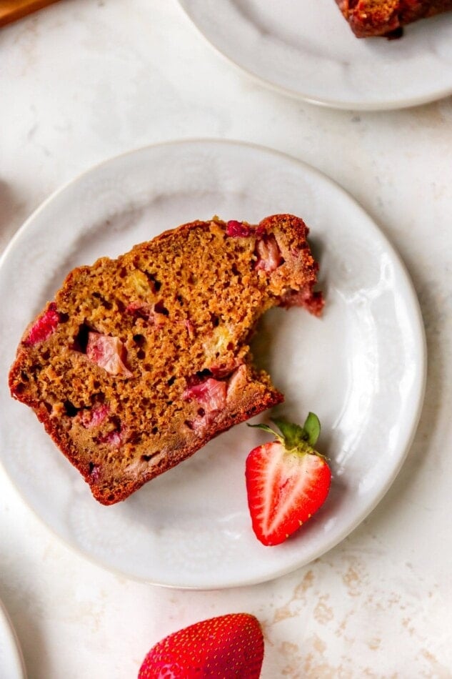A slice of strawberry banana bread with a bite taken out of it, resting on a plate with a halved strawberry.