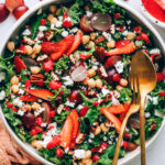 A serving bowl with strawberry kale salad. Serving utensils rest in the bowl.