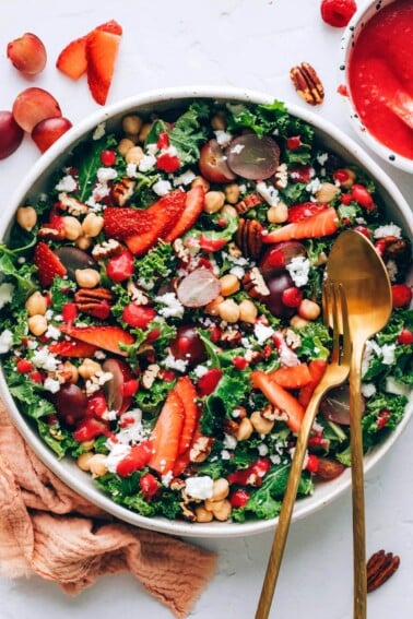 A serving bowl with strawberry kale salad. Serving utensils rest in the bowl.