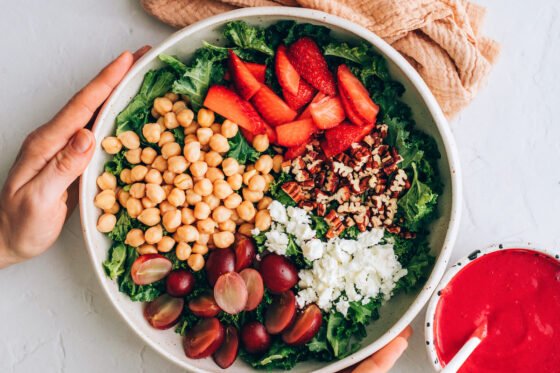 Ingredients for strawberry kale salad in a bowl together: kale, strawberries, grapes, chickpeas, crumbled feta and toasted pecans.