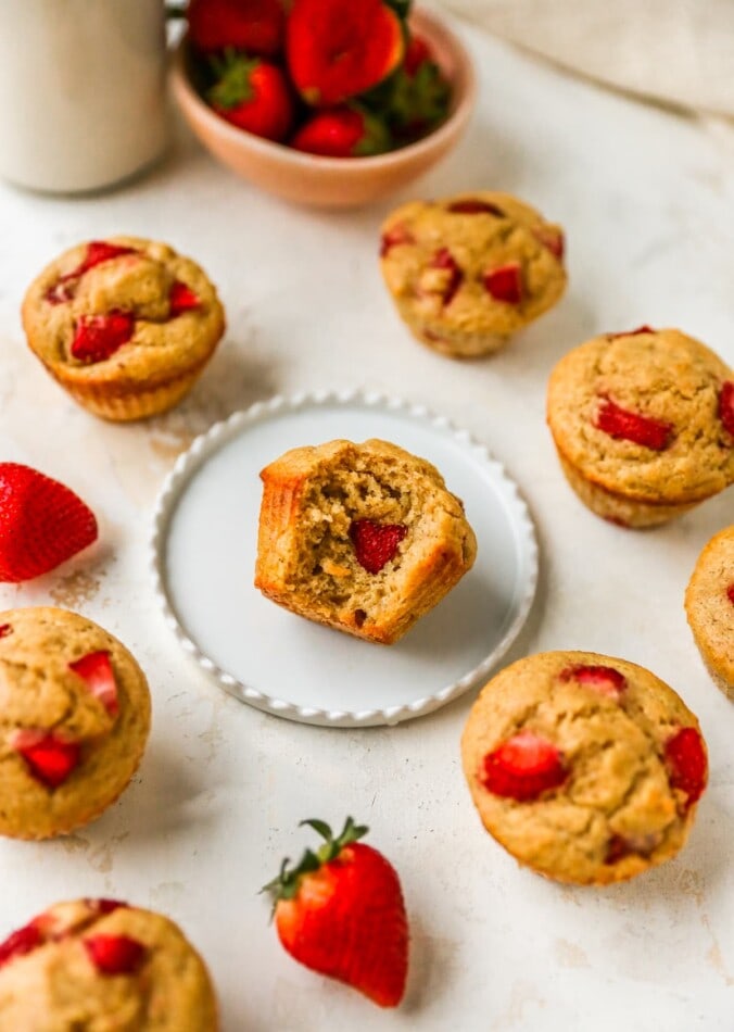 A muffin on a circle plate with a bite removed. The plate is surrounded by other muffins.