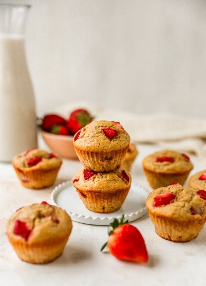 Two strawberry yogurt muffins stacked on top of each other on a plate. Other muffins are scattered around the plate.