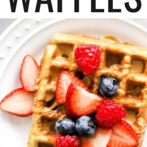 An almond flour waffles on a white plate. It has been topped with various berries and a drizzle of maple syrup. Fork has taken a bite of the waffle.