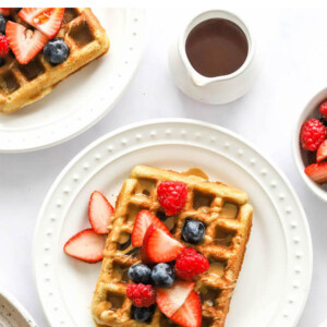 An almond flour waffles on a white plate. It has been topped with various berries and a drizzle of maple syrup.