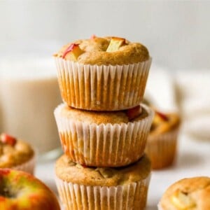 Three apple yogurt muffins stacked on top of each other. There are other muffins scattered around with apples.