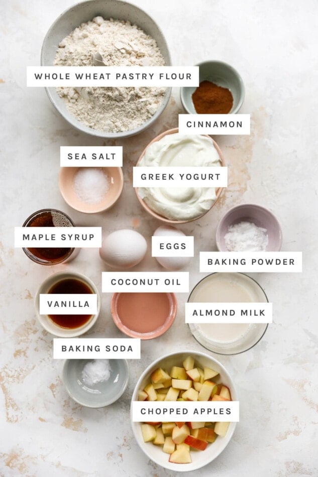 Ingredients measured out to make apple yogurt muffins: whole wheat pastry flour, cinnamon, Greek yogurt, sea salt, maple syrup, eggs, baking powder, coconut oil, vanilla, almond milk, baking soda and chopped apples.