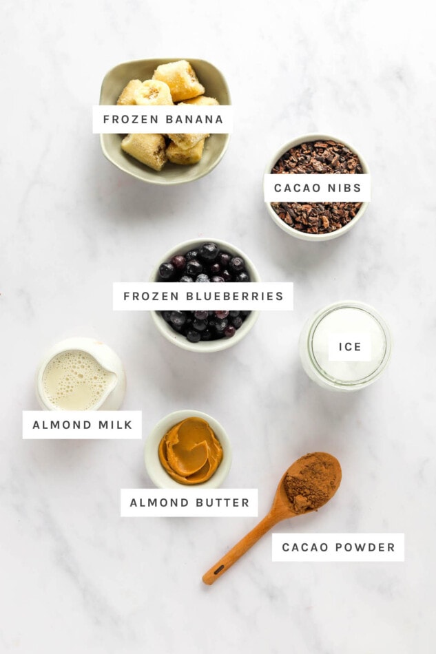 Ingredients measured out to make a cacao smoothie: frozen banana, cacao nibs, frozen blueberries, ice, almond milk, almond butter and cacao powder.