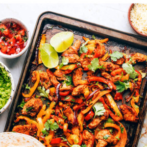 Chicken fajita filling on a sheet pan with a wooden spoon. Tortillas are resting on the bottom of the sheet pan.