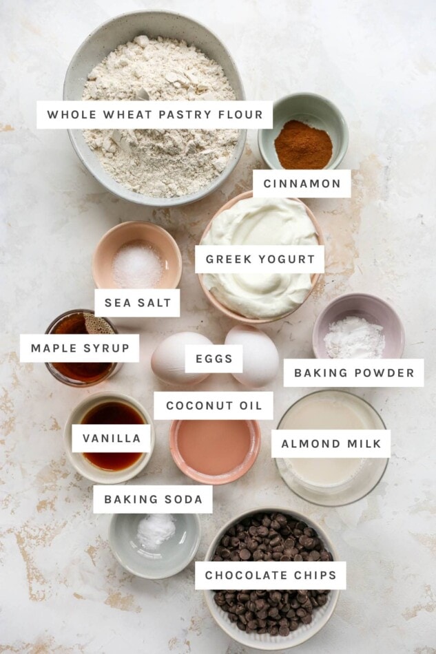 Ingredients measured out to make chocolate chip yogurt muffins: whole wheat pastry flour, cinnamon, Greek yogurt, sea salt, maple syrup, eggs, baking powder, coconut oil, vanilla, almond milk, baking soda and chocolate chips.