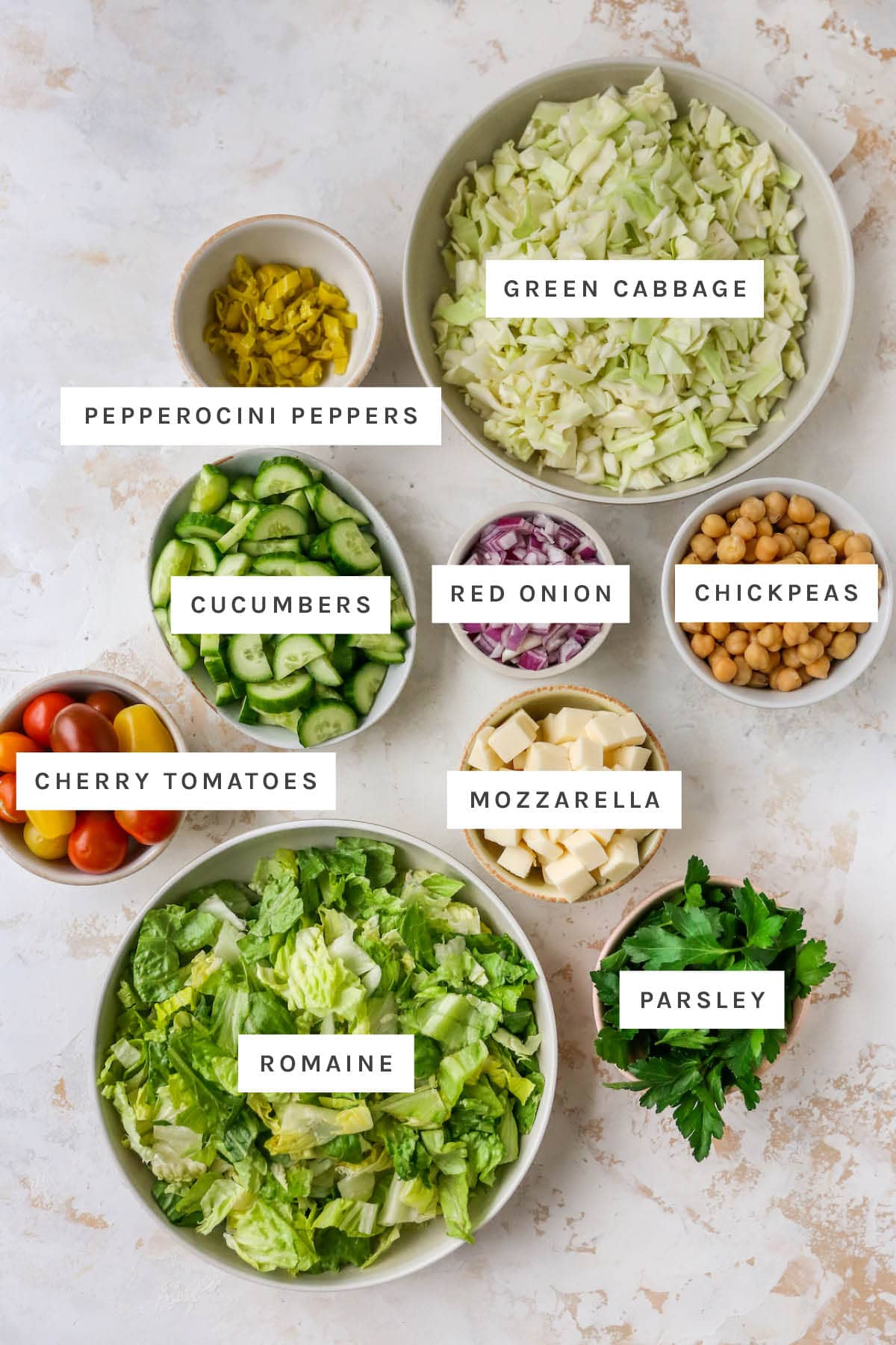 Ingredients measured out to make an Italian chopped salad: cabbage, pepperocini peppers, cucumber, red onion, chickpeas, mozzarella, cherry tomatoes, romaine and parsley.