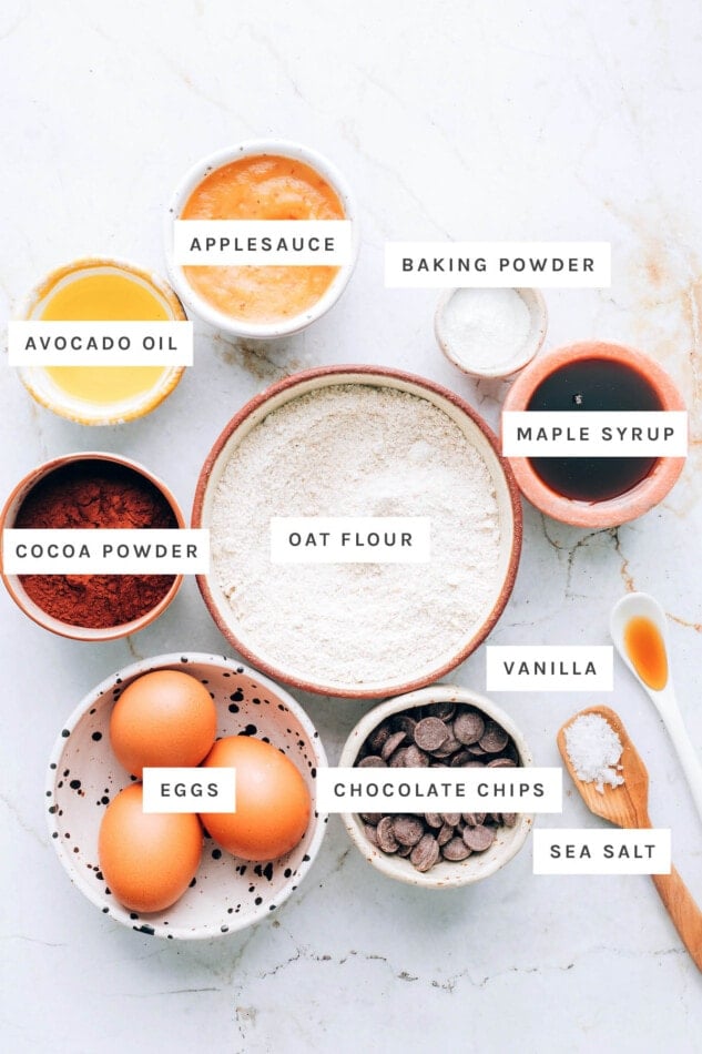 Ingredients measured out to make oat flour brownies: applesauce, avocado oil, baking powder, maple syrup, cocoa powder, oat flour, vanilla, eggs, chocolate chips and sea salt.