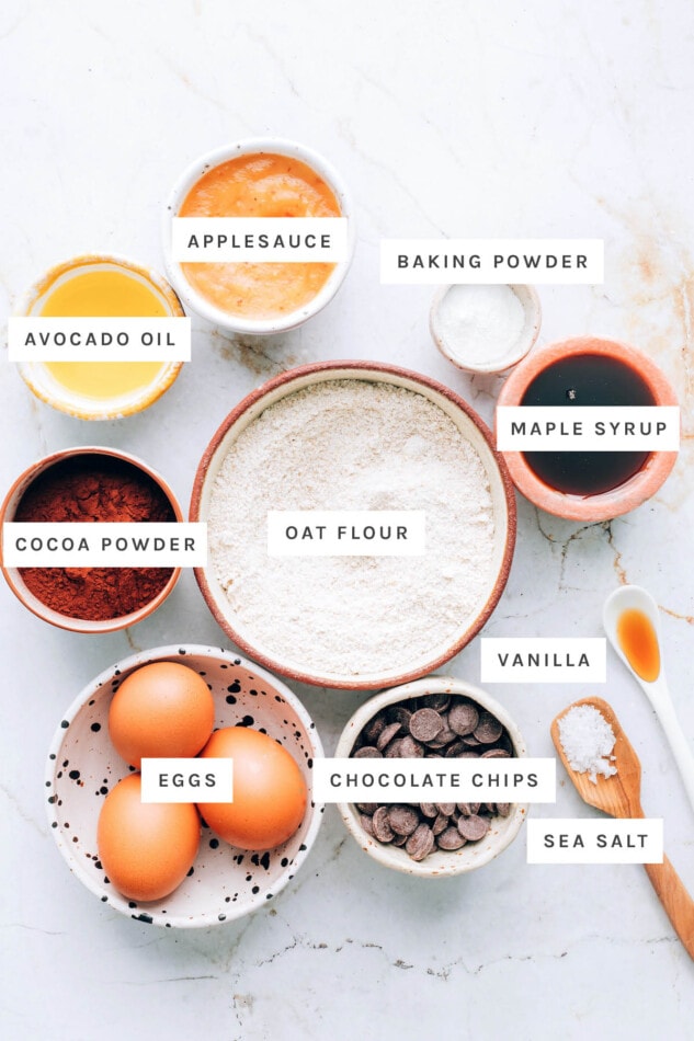 Ingredients measured out to make oat flour brownies: applesauce, avocado oil, baking powder, maple syrup, cocoa powder, oat flour, vanilla, eggs, chocolate chips and sea salt.