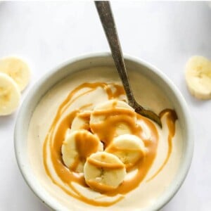 A small dish containing peanut butter protein pudding topped with banana slices and peanut butter drizzle. A spoon rests in the bowl.