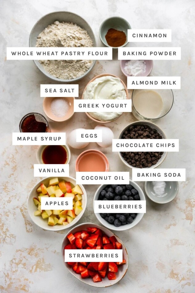 Ingredients measured out to make yogurt muffins 4 ways: whole wheat pastry flour, cinnamon, Greek yogurt, sea salt, maple syrup, eggs, baking powder, coconut oil, vanilla, almond milk, baking soda, strawberries, apples, blueberries and chocolate chips.