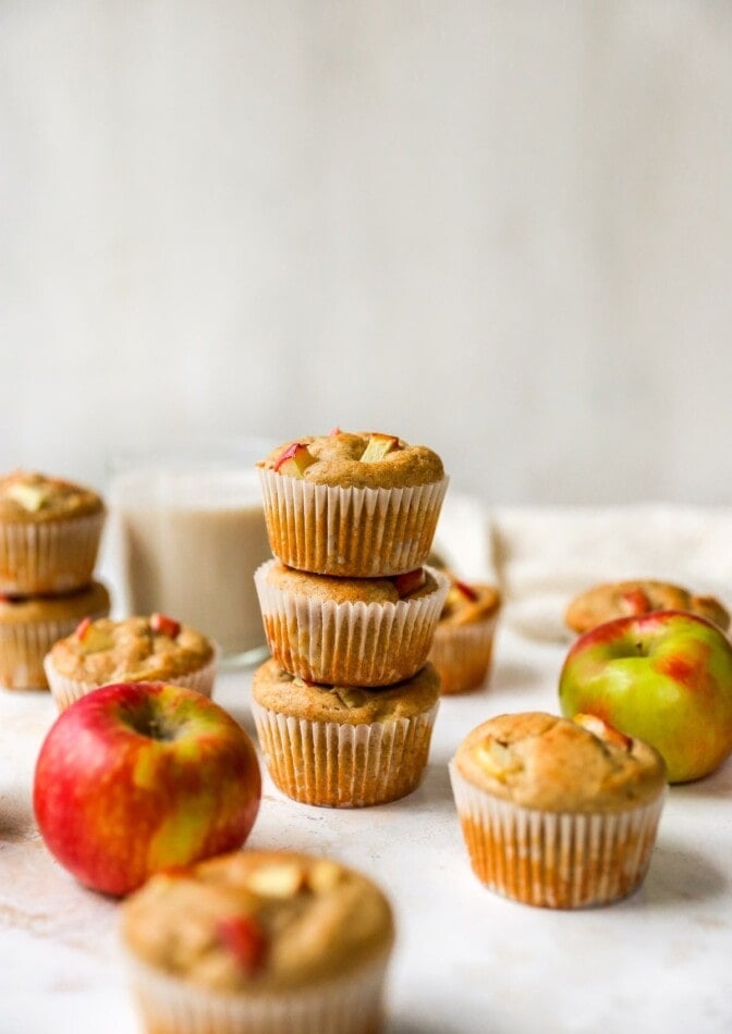 Three apple yogurt muffins stacked on top of each other. There are other muffins scattered around with apples.