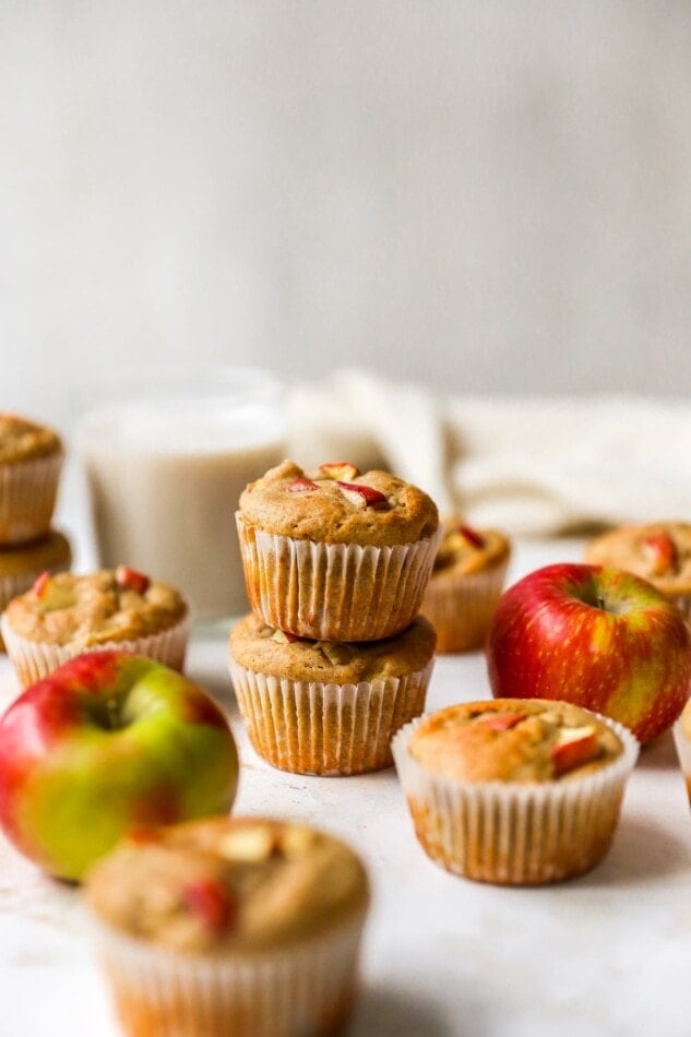 Two apple yogurt muffins stacked on top of each other. There are more muffins scattered around as well as whole apples.