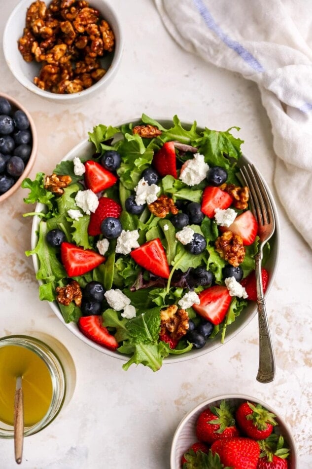 A bowl of summer berry salad with smaller bowls containing various toppings surrounding it.