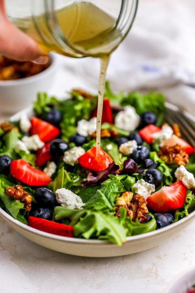 Pouring dressing from a mason jar onto a bowl of summer berry salad.