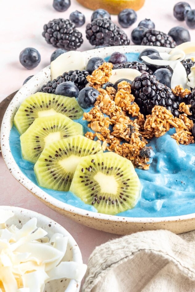 A blue smoothie in a bowl topped with kiwi, granola, blueberries, blackberries and coconut flakes.