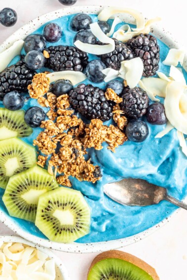 Overhead photo of a blue majik smoothie bowl topped with kiwi, granola, blackberries, blueberries and coconut flakes.