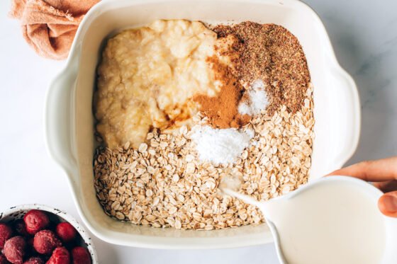 Oats, flaxseed, baking powder, cinnamon, salt, almond milk and almond extract in a baking dish. Almond milk is being poured into the dish.