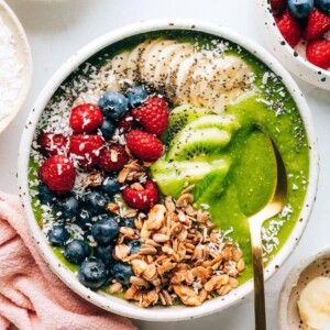 A green smoothie bowl topped with granola, shredded coconut, berries, banana and kiwi. A spoon rests in the bowl.