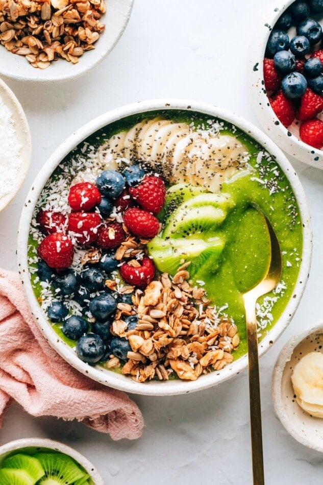 A green smoothie bowl topped with granola, shredded coconut, berries, banana and kiwi. A spoon rests in the bowl.