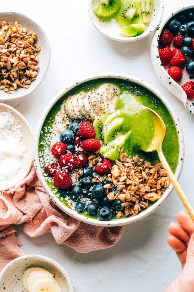 A green smoothie bowl topped with granola, shredded coconut, berries, banana and kiwi. A spoon is lifting out a bite.