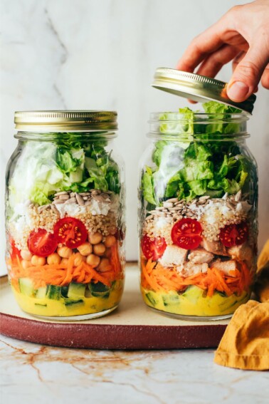 Two mason jar salads side by side, a hand is removing the lid of one jar.
