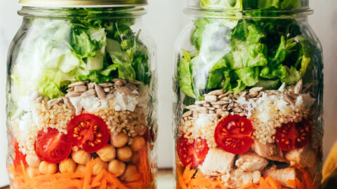 Two mason jar salads side by side, a hand is removing the lid of one jar.
