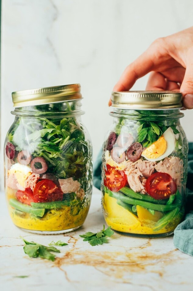 Two mason jars filled with Nicoise salad ingredients in layers. A hand is screwing on the lid to one of the jars.