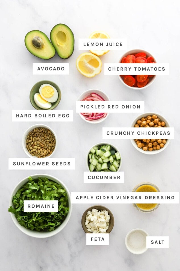 Ingredients measured out to make Mashed Avocado Bowl: avocado, lemon juice, cherry tomatoes, hard boiled egg, pickled red onion, crunchy chickpeas, sunflower seeds, cucumber, apple cider vinegar dressing, romaine, feta and salt.