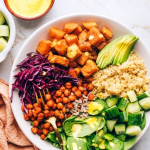 A buddha bowl with sweet potatoes, spinach, sunflower seeds, shredded red cabbage, chickpeas, cucumbers, quinoa and avocado. A fork rests in the bowl.