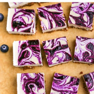 Blueberry swirled cheesecake bars on parchment paper surrounded by blueberries.