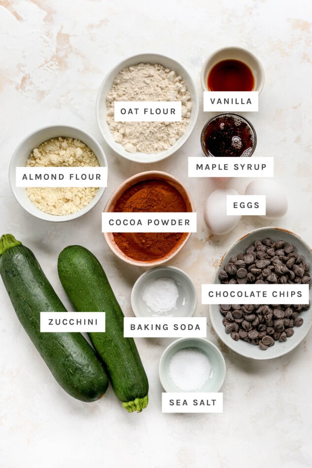 Ingredients measured out to make Zucchini Brownies: oat flour, vanilla, maple syrup, almond flour, eggs, cocoa powder, zucchini, baking soda, chocolate chips and sea salt.