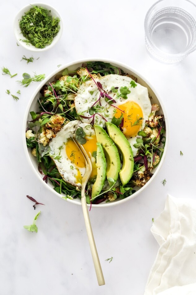 Overhead view of a breakfast salad, a fork is breaking the yolk of one of the eggs.