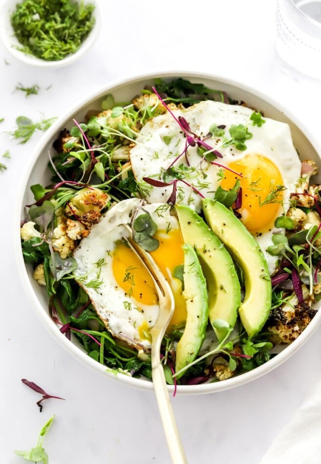 A breakfast salad bowl topped with eggs and avocado. A fork is breaking the yolk of one of the eggs.